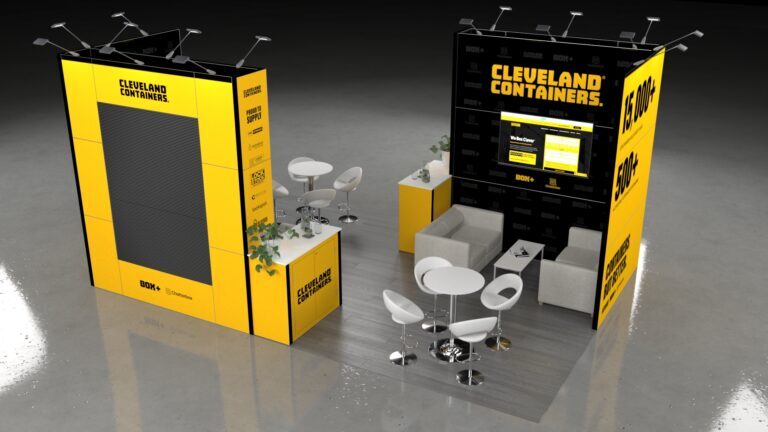 Large, open exhibition stand defined by two tall modular structures. The stand features one casual lounge area (sofa and armchair), a reception counter with storage, and a large wall graphic with an integrated digital screen.