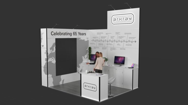 L-shaped exhibition stand featuring a reception counter and two workstations with computer monitors and under-counter storage. The stand has an elevated central back panel and large wall graphics.